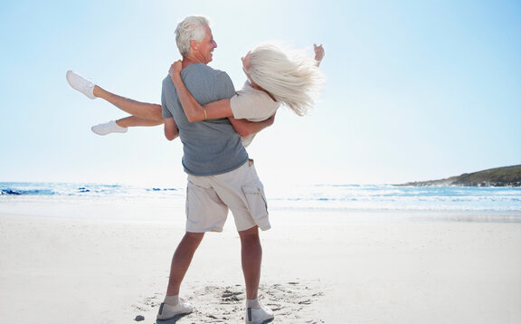 Glückliches Paar am Strand, älterer Mann trägt seine Frau dank starker Muskulatur mit Medizinischer EMS.