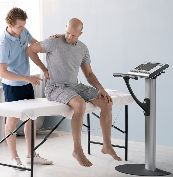 Mann mit Rückenschmerzen sitzt auf Behandlungsliege, daneben steht das Gerät für Medizinische EMS.